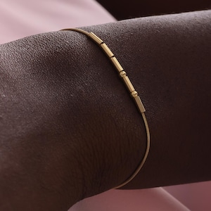 以下が含まれることがあります： 小さな長方形と円筒形のビーズが連なった繊細なゴールドブレスレット。 濃い色の腕に着用されており、背景はソフトピンクです。 ブレスレットの長さは約17〜20cmです。