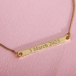 Puede incluir: Un collar dorado con una barra grabada con la fecha "3 March 2025". El colgante rectangular cuelga de una fina cadena. El fondo es de color rosa suave.