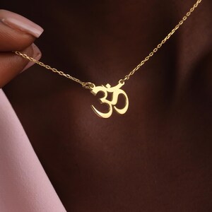 Könnte beinhalten: Eine goldfarbene Halskette mit einem Om-Symbol-Anhänger. Der Anhänger ist eine stilisierte Darstellung des heiligen Klanges. Die Halskette hat eine zarte Kette. Der Hintergrund ist unscharf und neutral.