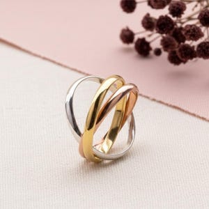 Könnte beinhalten: Ein Ring mit drei Bändern und ineinandergreifenden Bändern in Silber, Gold und Roségold. Die Bänder sind glatt und glänzend und schaffen ein modernes und elegantes Design. Der Ring ist vor einem weichen, neutralen Hintergrund platziert.