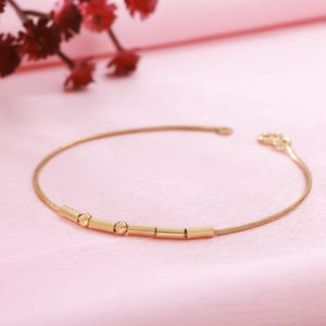 Könnte beinhalten: Ein zartes goldfarbenes Armband mit minimalistischem Design. Das Armband hat eine dünne, flexible Kette und ist mit kleinen, zylindrischen und kugelförmigen Goldperlen verziert. Das Armband wird auf einer rosa Oberfläche präsentiert.