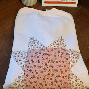 May include: A light pink crewneck sweatshirt featuring a large star-shaped appliqué on the front. The star is constructed from two different floral fabrics: one with small red flowers on a cream background and one with red flowers on a pink background.