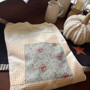 May include: A cream-colored sweatshirt featuring a central star-shaped appliqué. The star is constructed from a light green fabric with a small pink floral pattern, surrounded by points made of a tan and white gingham check fabric.