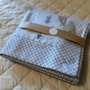 May include: Folded baby blanket with a brown paper band. The blanket features a white and gray grid pattern, with sections of light blue tree and boat designs. A white sticker with black text is visible on the band.