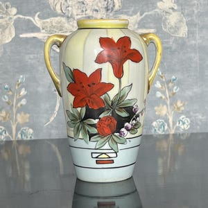 May include: A vintage ceramic vase with a light yellow and blue striped background. The vase features red floral designs and two gold handles. The base is light blue with geometric patterns. A decorative piece for home decor.