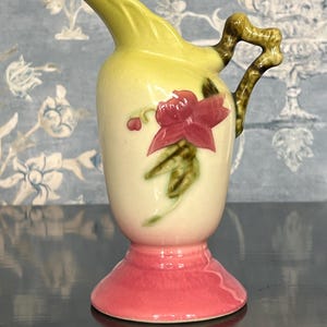 May include: A small, decorative pitcher with a yellow spout and handle. The body is cream-colored with a red floral design. The base is a vibrant pink. The handle has a brown, textured appearance. A charming piece for display.