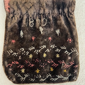 May include: A brown velvet drawstring pouch with embroidered details. The year "1870" is stitched on the front, along with floral and star patterns in white, pink, and yellow thread. The pouch has a pink lining.