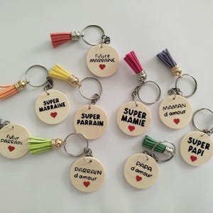 Peut inclure: Plusieurs porte-clés avec des pendentifs ronds en bois et des pompons colorés. Chaque pendentif porte une inscription différente en noir, comme "Super Mamie" et "Papa d'amour", avec un petit cœur rouge. Les pompons sont de différentes couleurs.