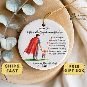 May include: A white ceramic ornament with a gold string, featuring a graphic of a father and child. Text reads "Super Dad, A Man With Superhuman Abilities" and lists abilities. The ornament is personalized with "Love you, Noah & Ally!" and the year 2025.