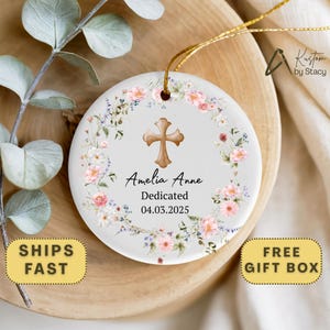 May include: Round ceramic ornament with a floral wreath design in pink and white. A brown cross is centered above the text "Amelia Anne Dedicated 04.03.2025". The ornament has a gold string for hanging.