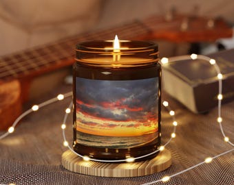 Puede incluir: Una vela encendida en un tarro de vidrio marrón con una etiqueta de escena de puesta de sol, colocada sobre un posavasos de madera. La vela está rodeada por una cadena de pequeñas luces blancas cálidas. Una guitarra y un libro están en el fondo.