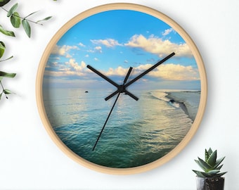May include: A round wall clock with a light wood frame. The clock face features a scenic view of a beach with blue water, a sandy shore, and a bright blue sky with fluffy white clouds. Black clock hands indicate the time.