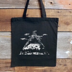 May include: Black canvas tote bag with a white graphic of a mountain range inside a triangle. The mountain range is snowy and has trees at the base.