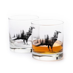 May include: Two clear whiskey glasses with a black and white raven and forest design. One glass is filled with amber liquid. The raven design is a silhouette of a bird with a forest scene inside.