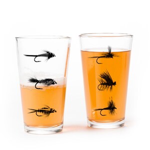 May include: Two clear glass pint glasses with a design of black fishing flies on the side. One glass is filled with a light orange liquid, the other is empty.
