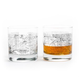 May include: Two clear, lowball glasses with a map design of Great Smoky Mountains National Park. One glass is filled with a golden liquid. The text "GREAT SMOKY MOUNTAINS NATIONAL PARK" is visible on the glass.