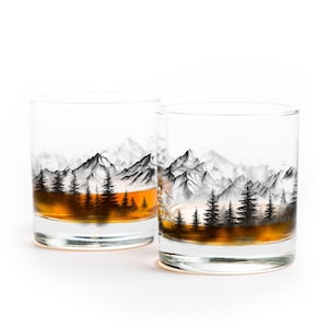 May include: Two clear whiskey glasses with a mountain and forest design. The glasses are filled with amber liquid. The mountain range is depicted in black and white, with a forest of trees at the base.