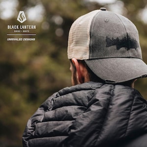 May include: A person wearing a grey and beige trucker hat with a black silhouette of a forest on the front. The hat has a distressed look and the text "BLACK LANTERN" and "ROCKS - ROOTS" is visible on the front of the hat.