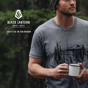 May include: A gray t-shirt with a black and white graphic of a mountain scene. The shirt has the text "BLACK LANTERN ROCKS + ROOTS CRAFTED IN COLORADO" on the front.