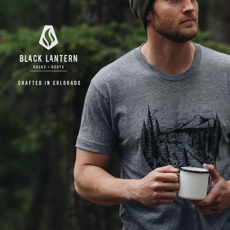 May include: A gray t-shirt with a black and white graphic of a mountain scene. The shirt has the text "BLACK LANTERN ROCKS + ROOTS CRAFTED IN COLORADO" on the front.