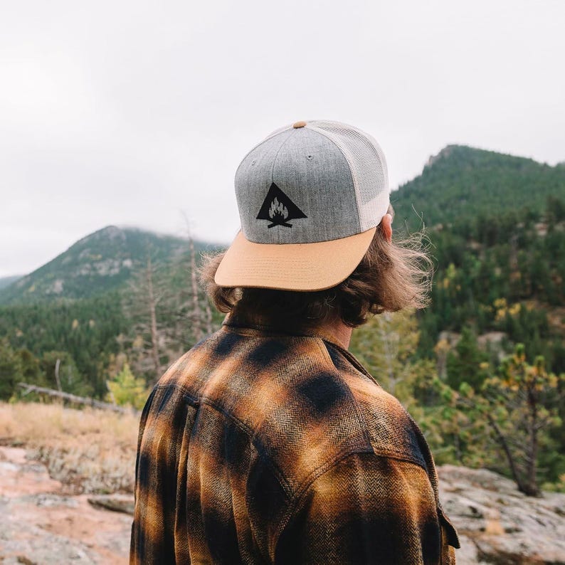 May include: A person wearing a grey and tan baseball cap with a black campfire logo. The cap has a mesh back. The person is wearing a plaid shirt and is standing in front of a mountain range.