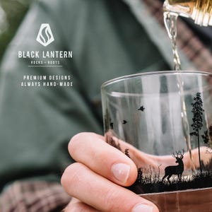 May include: A clear glass tumbler with a black forest scene featuring a deer, trees, and birds. The glass is being filled with a golden liquid. The tumbler has the text "BLACK LANTERN" and "ROCKS + ROOTS" printed on it.