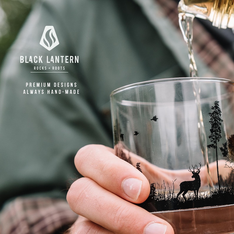 May include: A clear glass with a black and white design of a deer in a forest setting. The glass is being filled with a golden liquid. The text "BLACK LANTERN ROCKS + ROOTS PREMIUM DESIGNS ALWAYS HAND-MADE" is visible in the background.