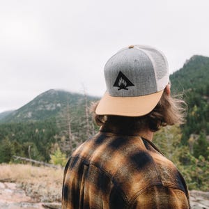 May include: A person wearing a gray and tan baseball cap with a black campfire logo. The cap has a mesh back. The person is wearing a plaid shirt and is standing in front of a mountain range.