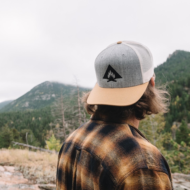 May include: A person wearing a gray and tan baseball cap with a black campfire logo. The cap has a mesh back. The person is wearing a plaid shirt and is standing in front of a mountain range.