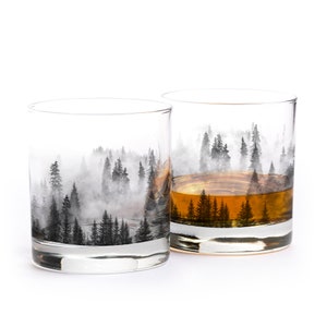 May include: Two clear whiskey glasses with a forest scene design. The glasses feature a black and white illustration of a misty forest. One glass is filled with amber liquid, the other is empty.