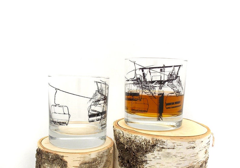 Two ski lift whiskey tumbler glasses sitting one each atop two different small pieces of aspen wood cut into different height sizes.