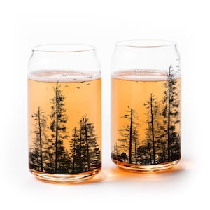 May include: Two clear glass drinking glasses with a forest silhouette design. The glasses are filled with a light orange beverage.