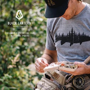 Pu&ograve; includere: Un uomo indossa una maglietta grigia con una silhouette nera di un pesce e alberi stampati su di essa. La maglietta ha il testo "Black Lantern Rocks + Roots" e "Proudly Supporting Our National Parks". L'uomo tiene una scatola da pesca a mosca con diverse mosche.