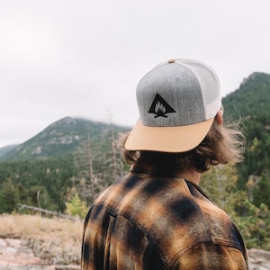 May include: Gray and tan trucker hat with a black triangle logo featuring a campfire design. The hat has a mesh back and a tan brim. The person in the image is wearing a brown and black plaid shirt.