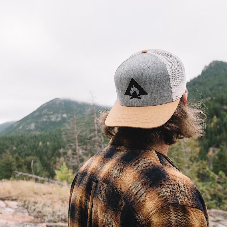 May include: Gray and tan trucker hat with a black triangle logo featuring a campfire design. The hat has a mesh back and a tan brim. The person in the image is wearing a brown and black plaid shirt.