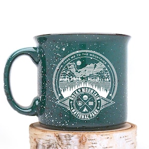 May include: Green speckled ceramic mug with a white graphic of a mountain scene and the text "Take me to the mountains" and "Rocky Mountain National Park".