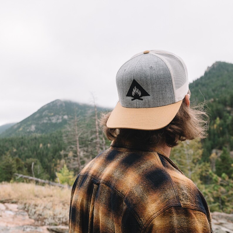 May include: A person wearing a gray and tan baseball cap with a black campfire logo. The cap has a mesh back. The person is wearing a plaid shirt and is looking out at a mountain range in the distance.