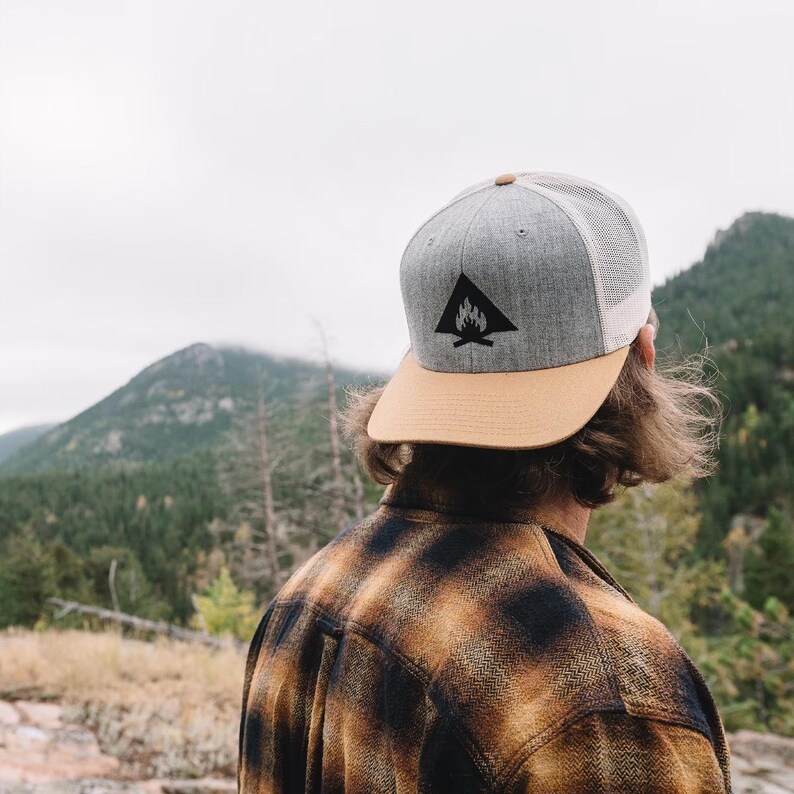 May include: A person wearing a gray and tan baseball cap with a black campfire logo. The cap has a mesh back and a tan brim. The person is wearing a plaid shirt and is standing in front of a mountain range.