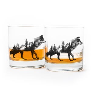 May include: Two clear whiskey glasses with a black and white wolf and forest design. The glasses are filled with amber-colored liquid. The wolf design is a silhouette of a wolf walking through a forest.