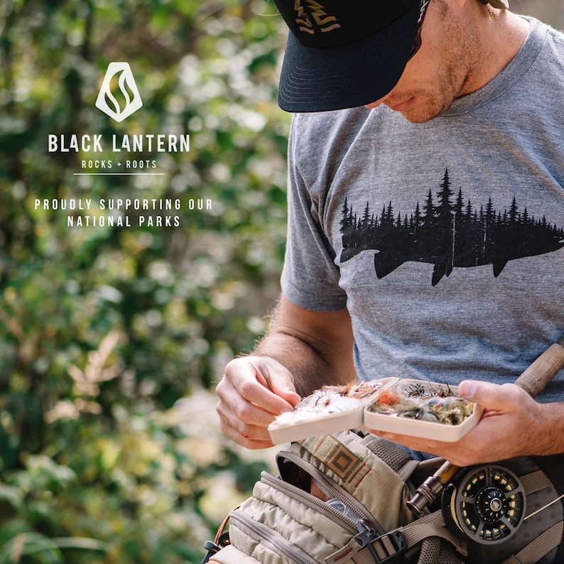 May include: A person wearing a grey t-shirt with a black fish and trees graphic, a black baseball cap, and a brown and beige fishing vest. The t-shirt has the text "Black Lantern Rocks + Roots" and "Proudly Supporting Our National Parks".