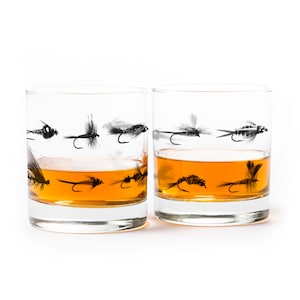 May include: Two clear glass tumblers with a pattern of black fly fishing lures around the bottom. The tumblers are filled with amber liquid.