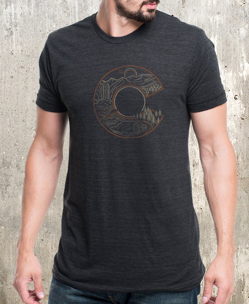 May include: Dark gray t-shirt featuring a brown outline of the letter "C" with a landscape design inside. The design includes mountains, a waterfall, trees, and a lake. The shirt is made of a soft material.