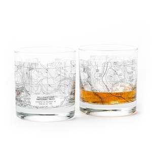 May include: Two clear, cylindrical glasses with a map of Yellowstone National Park printed on them. One glass is filled with a golden liquid. The text "YELLOWSTONE NATIONAL PARK" is visible on the glass, along with coordinates and other details.
