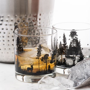May include: Two clear glass tumblers with black and white forest silhouette designs. The tumblers are filled with ice and a brown liquid. One tumbler has a deer silhouette in the foreground.