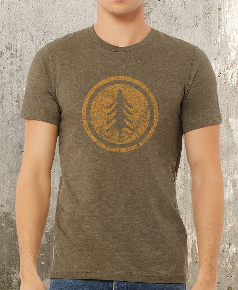 Retro map tree graphic print design in golden yellow ink shown in the center of the heather green t shirt which is on a person shown between the neck and hips so you see the exact fit of the t-shirt.