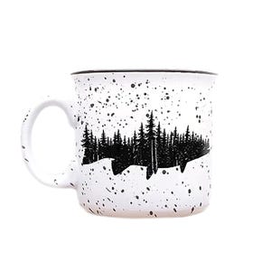 May include: White ceramic mug with a black silhouette of a fish and a forest line. The mug has a speckled white glaze.