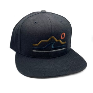 Könnte beinhalten: Marineblaue Baseballkappe mit flachem Schirm und gesticktem Design einer Bergkette, Wellen und einer untergehenden Sonne in Gold, Blau und Orange. Die Kappe hat eine strukturierte Krone und einen gebogenen Schirm.