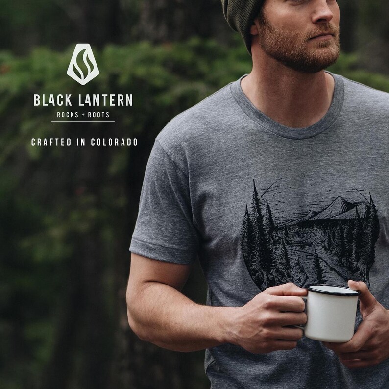 May include: A gray t-shirt with a black graphic of a mountain scene and the text "BLACK LANTERN ROCKS + ROOTS CRAFTED IN COLORADO." A white enamel mug with a black rim is held in the hands.