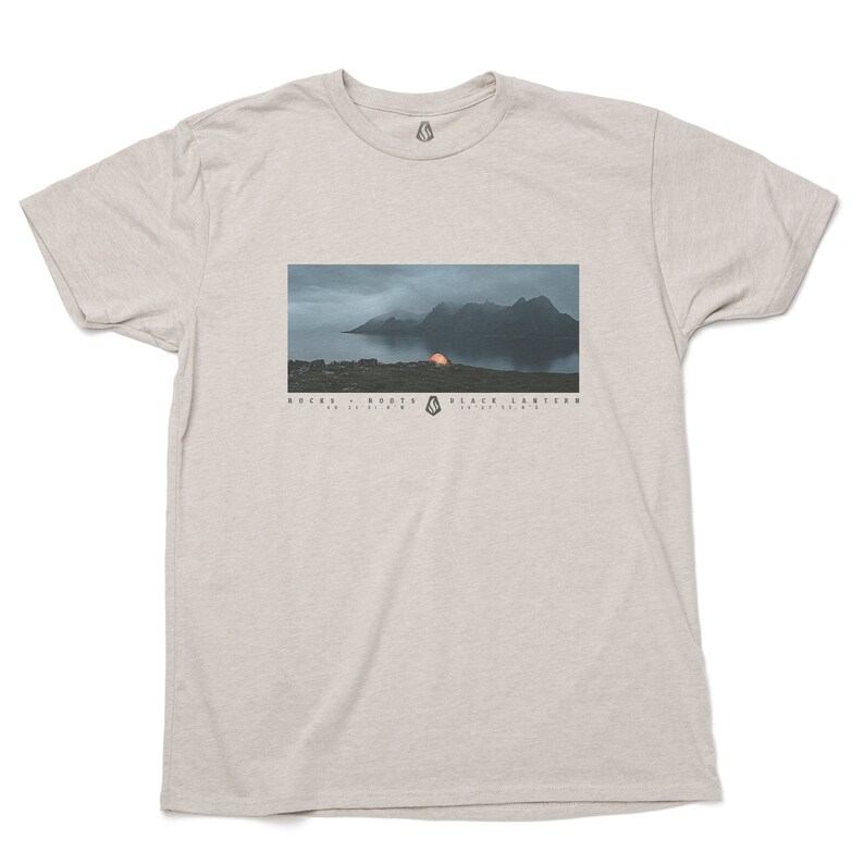 May include: A heather grey t-shirt with a graphic of a mountain range and a tent with a glowing orange light. The text "ROCKS - ROOTS - BLACK LANTERS" is printed below the image.