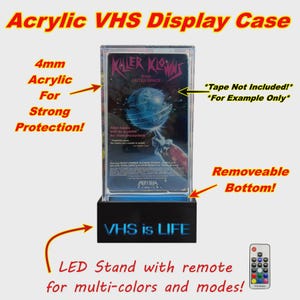 Könnte beinhalten: Transparente Acryl-VHS-Display-Hülle mit abnehmbarem Boden. Die Hülle verfügt über einen schwarzen LED-Ständer mit dem Text "VHS is LIFE" und eine Fernbedienung für Mehrfarbmodi. Starker Schutz.